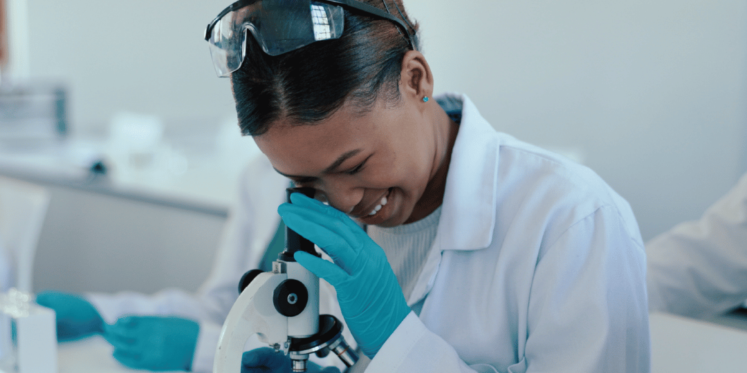 Young, female learner looking through a monocular microscope wearing protective glass, a white lab coat and blue gloves.