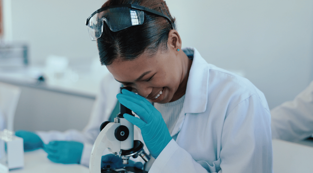Young, female learner looking through a monocular microscope wearing protective glass, a white lab coat and blue gloves.