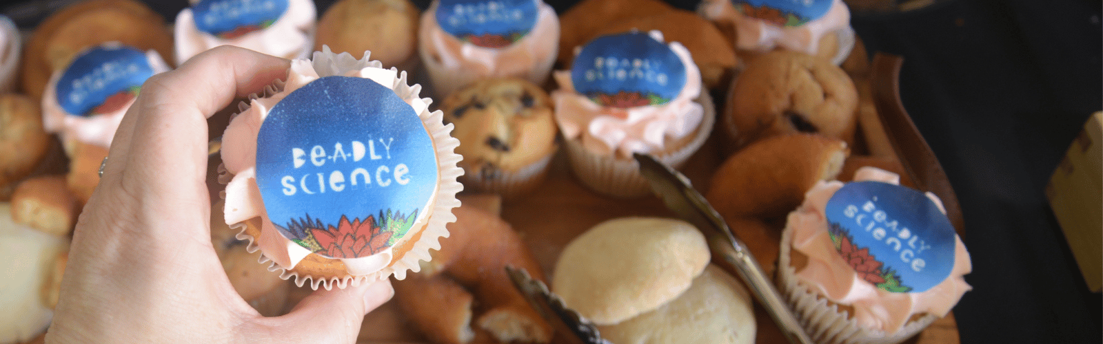Cupcake with 'DeadlyScience' icing image held by a hand, surrounded by other cupcakes.