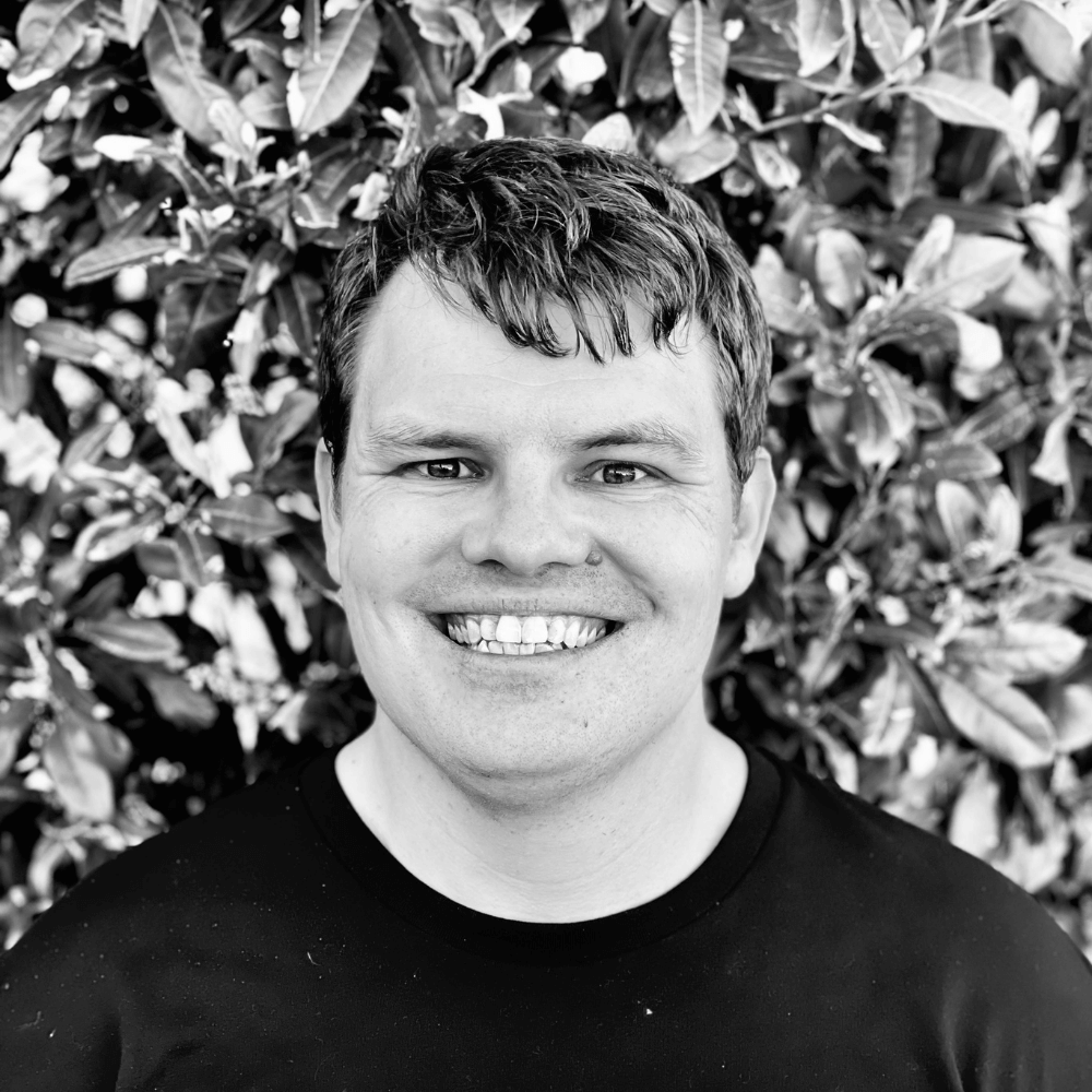 Black and white photo of Kamilaroi man, DeadlyScience, Founder and CEO, Corey Tutt. He is wearing a black t-shirt with a blurred natural background. 
