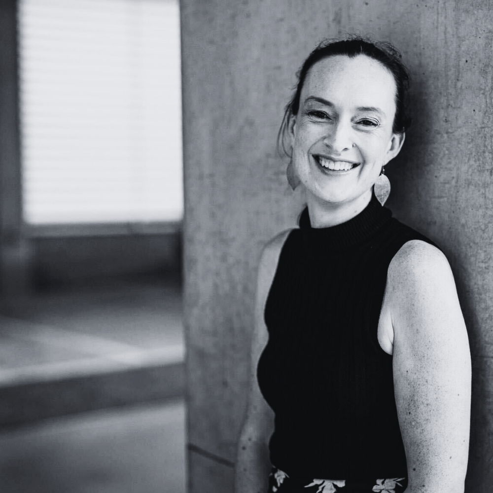 Black and white photo of a Rachel Peek standing against a textured wall, smiling at the camera.